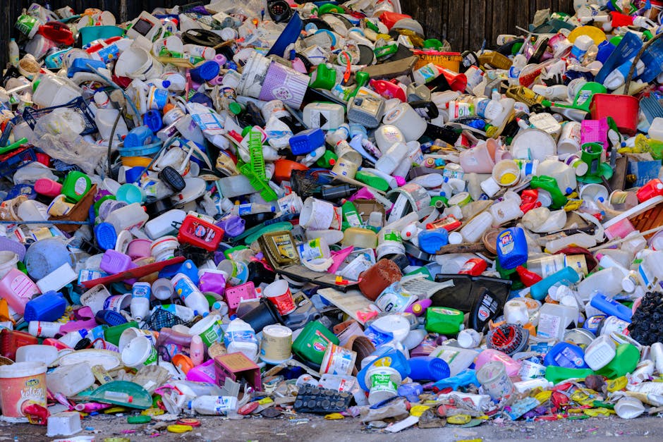 A large outdoor pile of mixed household rubbish includes numerous plastic containers, bottles, and cups in various colors such as white, blue, green, and pink, with some featuring labels and logos. Th
