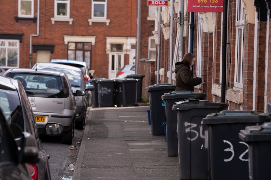 A middle-aged man with dark hair and light skin is seen on a paved urban street, bent over a metal litter bin while placing waste into the opening. He is wearing a dark T-shirt with a graphic print on